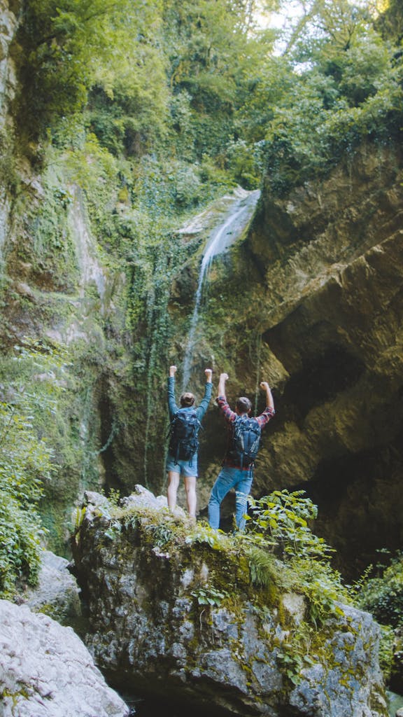 A couple of hikers raise their arms victoriously on a mossy rock near a scenic waterfall in a lush forest.
