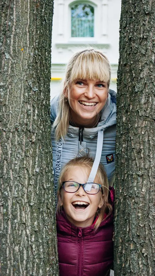 Happy mother and daughter smiling while posing between tree trunks outdoors.